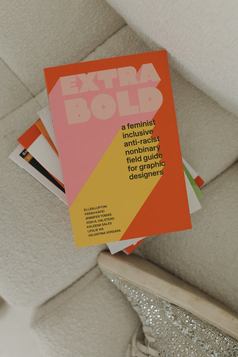 Book stack with the book "Extra Bold: A feminist inclusive anti-racist nonbinary field guide for graphic designers," on a couch next to a silver rhinestone shoe